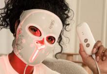 Close up image of person with black curly hair and a white top, wearing a white red-light therapy mask with red light illuminating from inside it. The mask says 'Silk'n' on the front, and the person is also holding up a white hand held device which controls the mask.