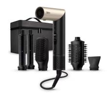 Image of hair styling set in black and gold, with two curling attachments, a paddle brush, a round brush and a concentrator nozzle alongside the main hair dryer unit. The attachments and unit are on a white background in front of a back travel case for the set. 