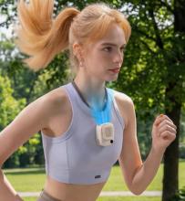 Image of woman jogging outdoors wearing a light purple sports top and a small white wearable neck fan attached to a lanyard around her neck. The fan is emitting a blue colour from the top to represent its neck cooling abilities. 