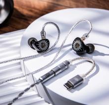Image of black in ear monitors with silver twisted cable, with a USB-C connector at one end. The IEMs are on a white circular platform. 