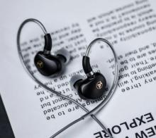 Image of black in-ear monitors with silver cable, on a white background with black text which is slightly blurred on it. 