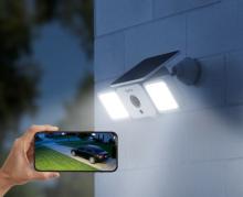 Image of a solar powered outdoor security light with two bright lights on either side mounted on an outside wall. The image also includes a hand holding a smartphone displaying a live feed image of a driveway and parked car. 