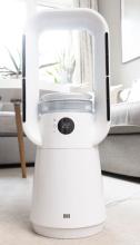 Image of white bladeless fan that has a rectangular loop design, with a black digital display on the front and air panels on each side. The background of the image is beige/grey living room setting. 