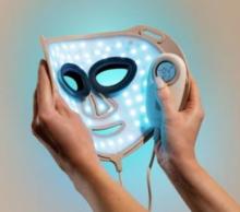 Image of hands holding up a light therapy face mask which is illuminated with blue LED lights, and has eye and mouth cutouts. One of the hands is also holding a small white device to control the mask. 